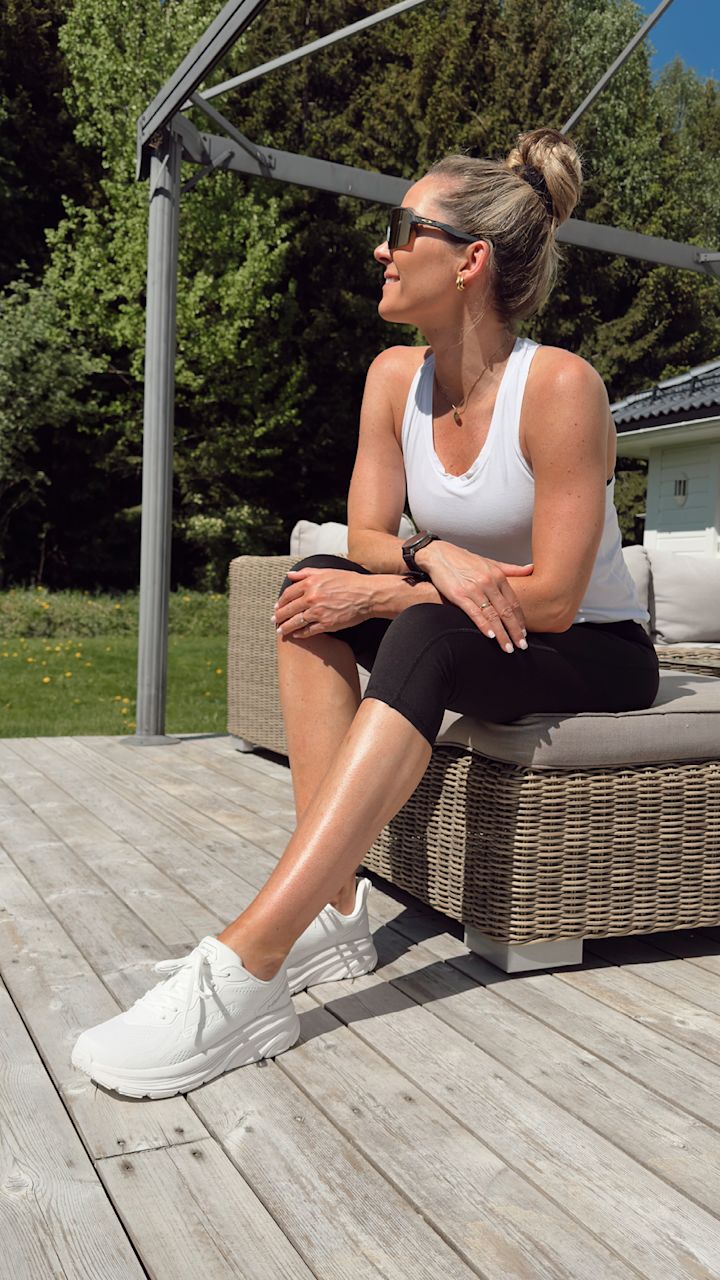 Kvinna i vit träningslinne, svarta träningsbyxor och vita joggingskor sitter på en flätad trädgårdsbänk på en träaltan utomhus med en grön trädgård och träd i bakgrunden.