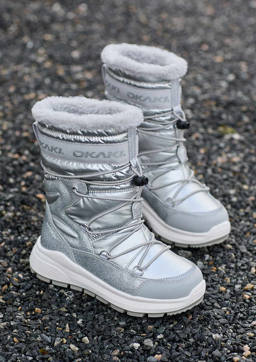 Silverfärgade vinterkängor från OKAKI med vit pälskant, snörning och glittrande detaljer, placerade på mörk grusyta.
