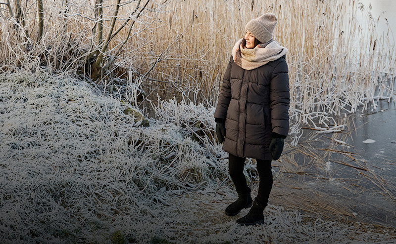 Kvinna klädd i vinterjacka, mössa, halsduk och vantar står på frosttäckt mark vid en isbelagd sjö med torrt vass i bakgrunden.