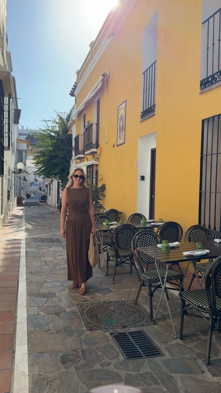 Kvinna i brun maxiklänning och bruna sandaler promenerar på en solig gata i Spanien med gula byggnader.