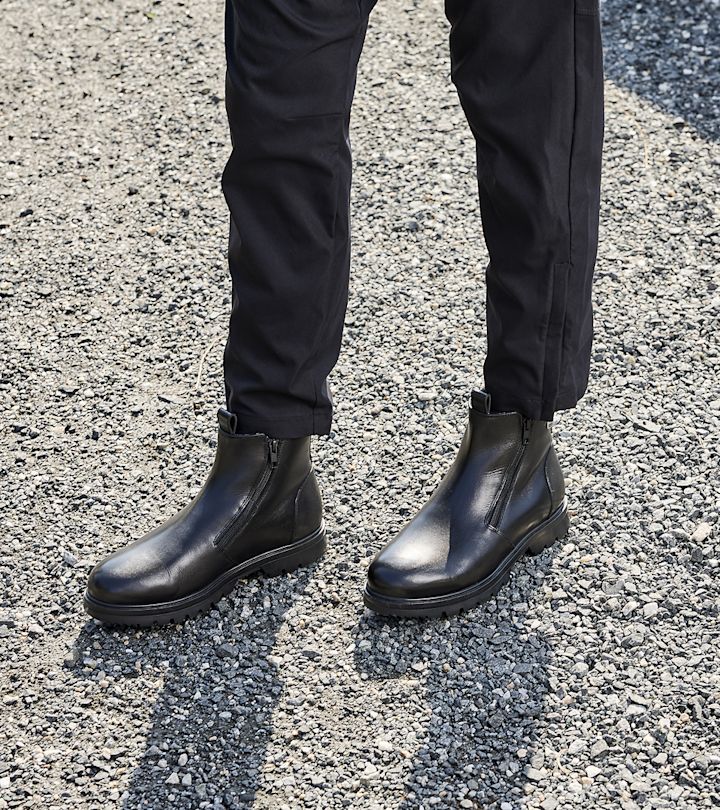 Svarta skinnstövlar med dragkedja på sidan, bärna av en person i svarta byxor, stående på grusbelagd mark.