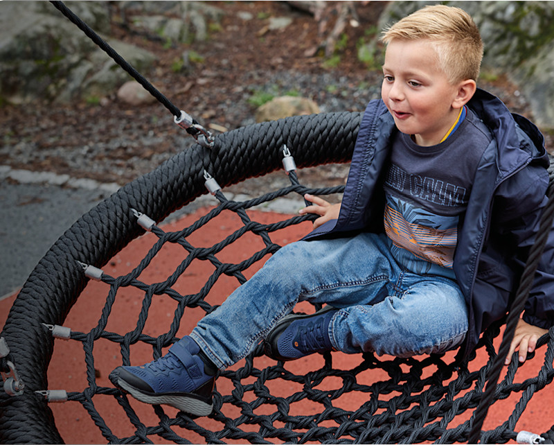 Barn i blå jacka och jeans sitter i nätgunga på lekplats, bär mörkblå övergångsskor från Okaki med kardborrespännen.