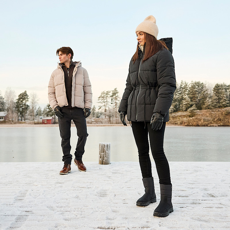 To personer kledd i vinterjakker, hansker og støvler står på en snødekt brygge ved en innsjø med trær i bakgrunnen.