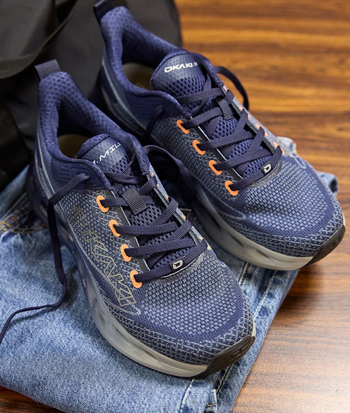 Mörkblå fritidsskor från Okaki med orange snörhål och nätmaterial, placerade på jeans på ett träbord.