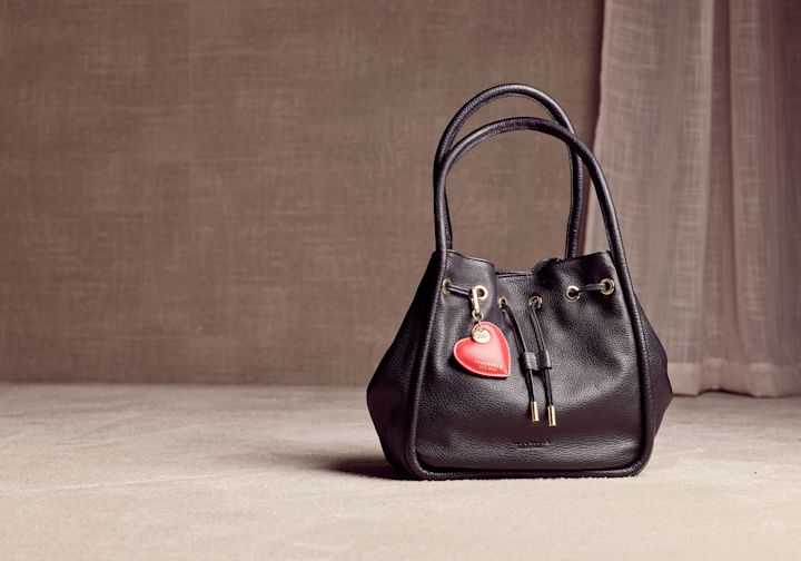 Elegant svart skinnveske med snøring og et rødt hjerteformet anheng, plassert mot en nøytral beige bakgrunn.