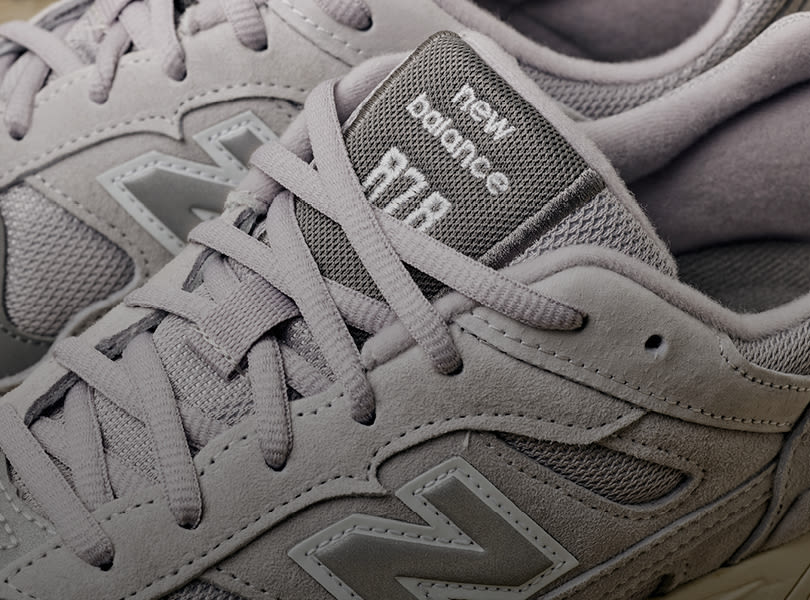 Grå New Balance joggesko i sporty stil med mesh og semsket skinn, vist i nærbilde med merkets logo på tungelappen.