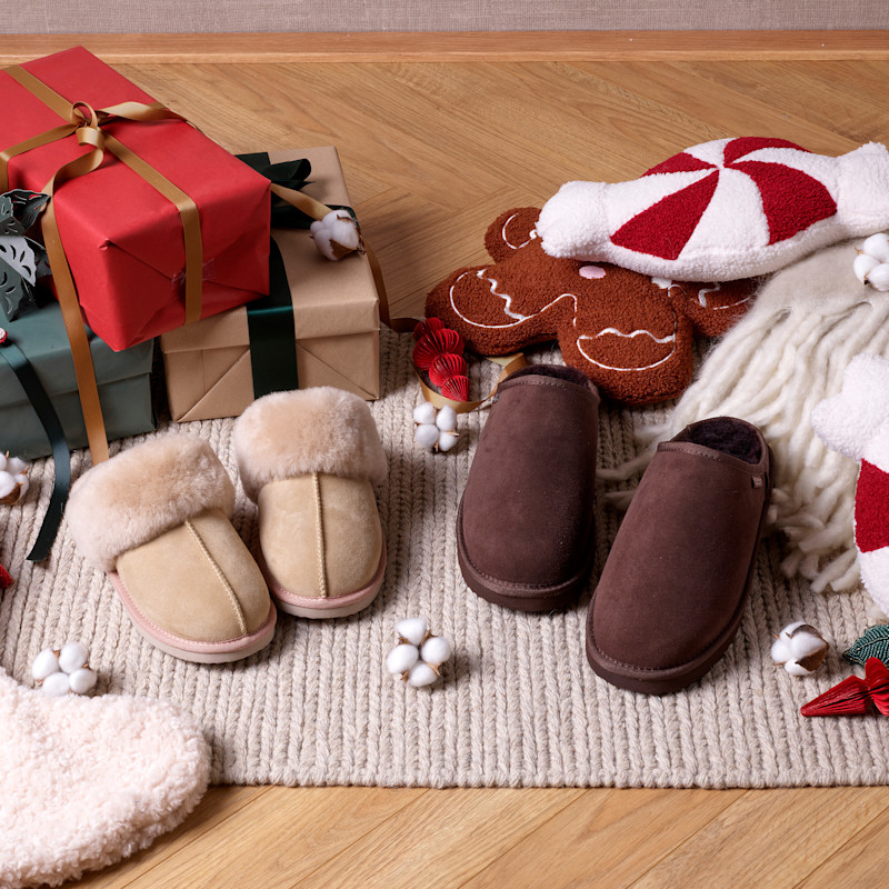 Beige och bruna mockaskor med pälskant bredvid julklappar och pepparkakskudde på en stickad matta.