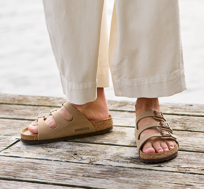 Beige Birkenstock sandaler med spænder og korksål, båret med cremefarvede bukser på en træterrasse ved vandet.