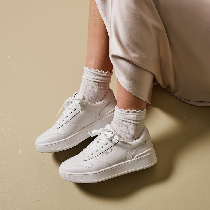 Hvite sneakers med tekst "STOCKHOLM DESIGN GROUP" på siden, iført med hvite sokker med bølget kant, på beige gulv ved en person med lys beige skjørt.
