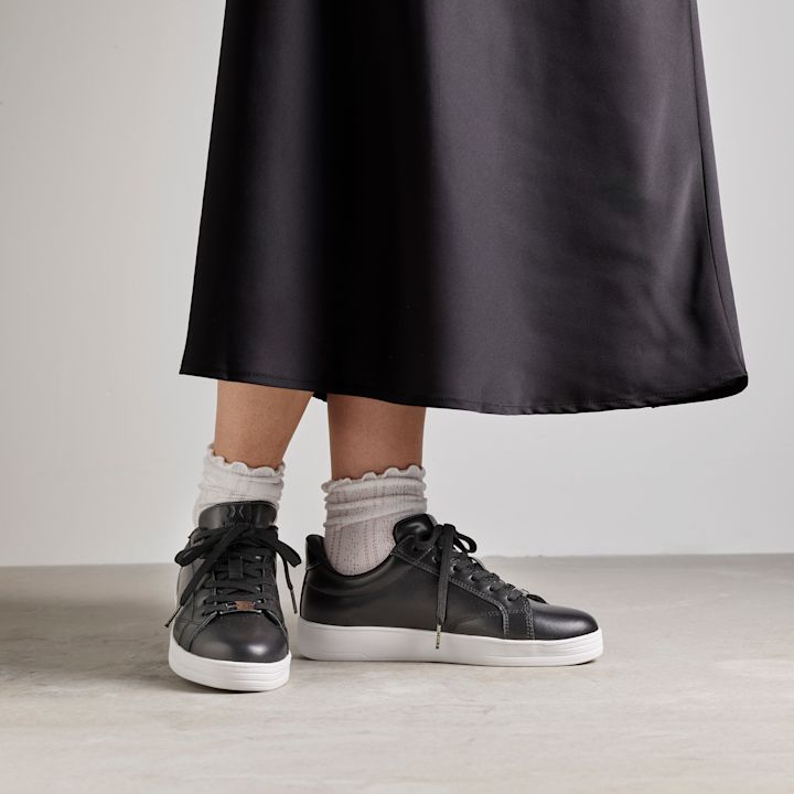 Svarta lädersneakers med vit sula, burna med vita volangstrumpor under en svart kjol mot en ljusgrå bakgrund.