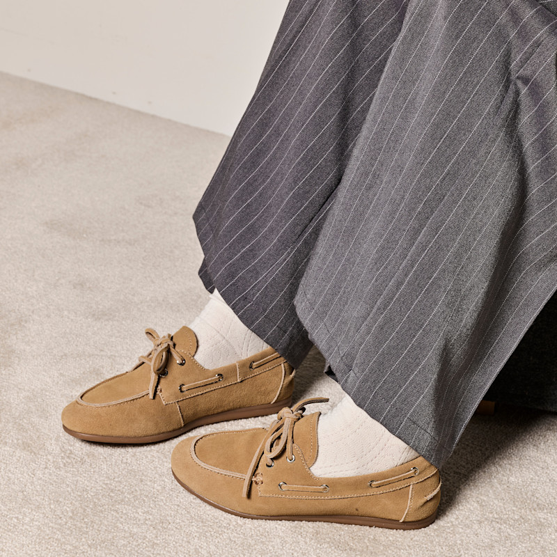 Beige mockasiner i semskskinnsliknande material med snörning, burna med vita strumpor och grårandiga byxor på ljus matta.