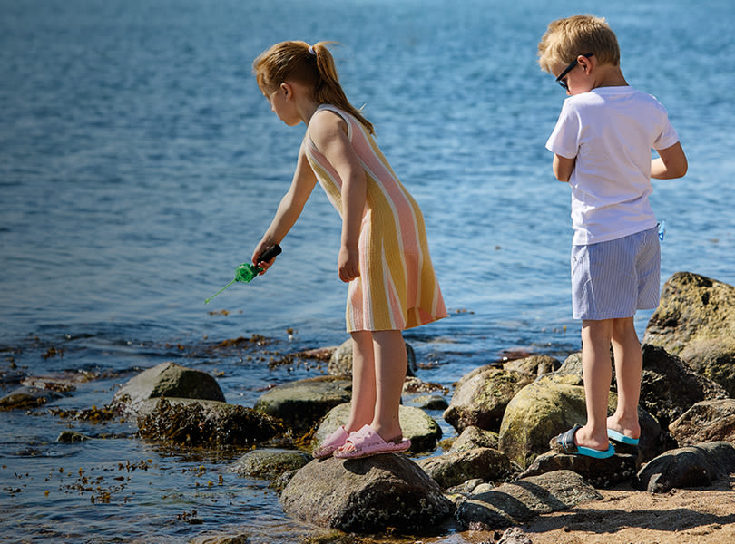 To barn utforsker stranden, jente med rosa sandaler og gutt med turkise sandaler står på steiner ved vannet på en solrik dag.