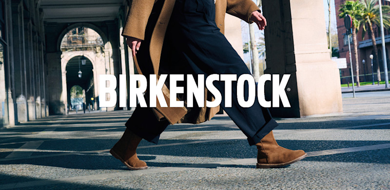 Person i kamelfarget kåpe og mørke bukser går med brune Birkenstock ankelstøvletter i semsket skinn på bygate.