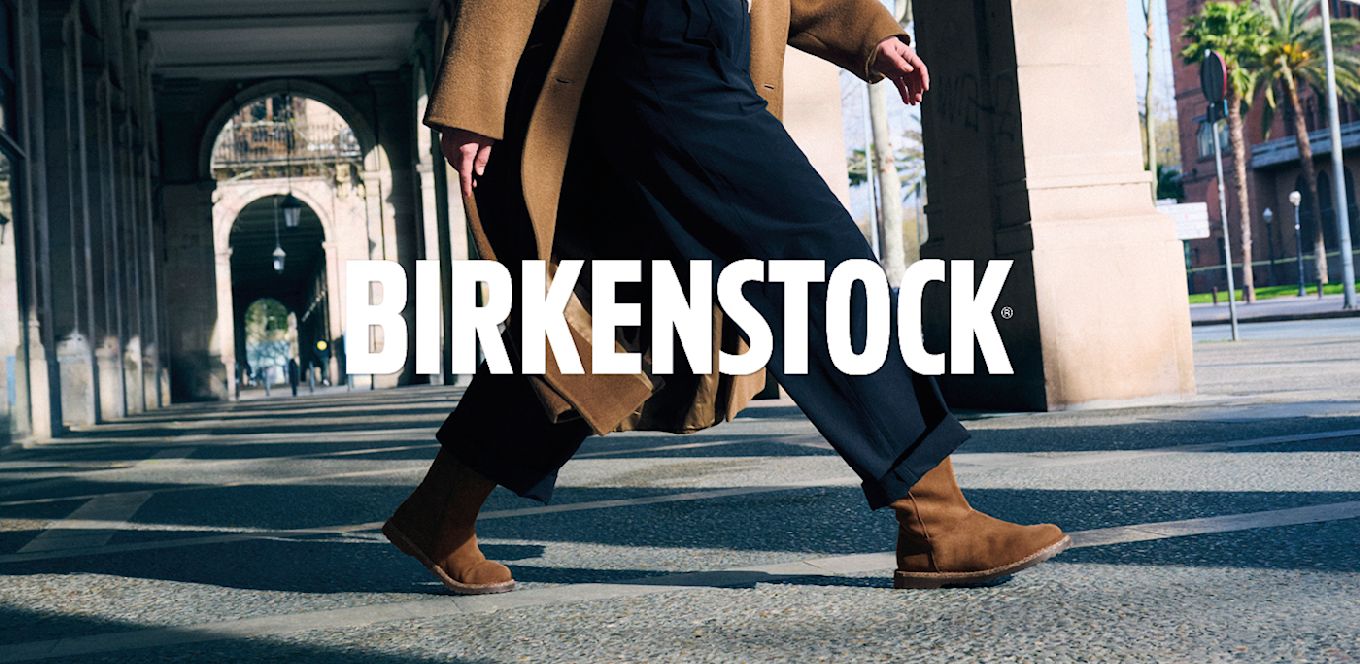 Person i beige kappa och svarta byxor går i bruna mockaboots från Birkenstock på stadsgata med arkader och palmer.