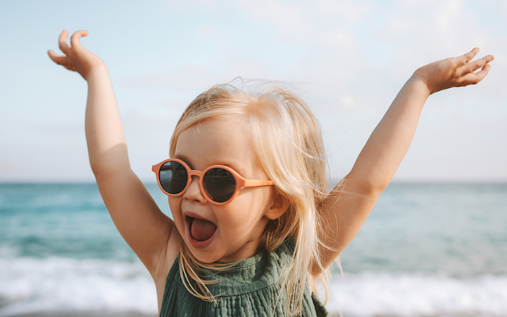 Lite barn med oransje solbriller og grønn kjole løfter armene og jubler ved stranden med havet i bakgrunnen.
