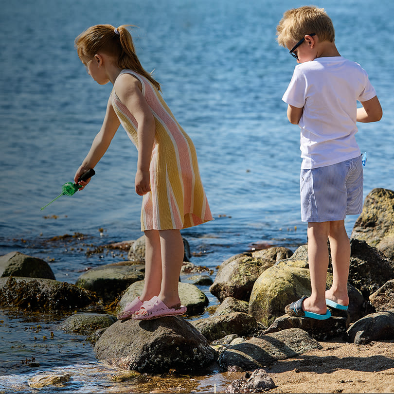 Två barn vid havet på klippor, flickan i rosa sandaler och pojken i turkosa vattenskor utforskar strandkanten.