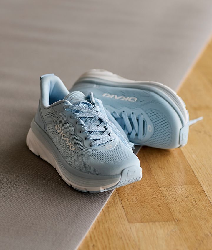 Lyseblå Okaki Hyper joggesko med nettingmønster og hvit såle, plassert på et grått teppe ved siden av tregulv.