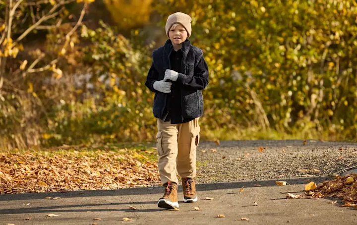 Barn i höstlandskap med beige mössa, mörk jacka och bruna snörkängor från Okaki promenerar på en stig omgiven av höstlöv.