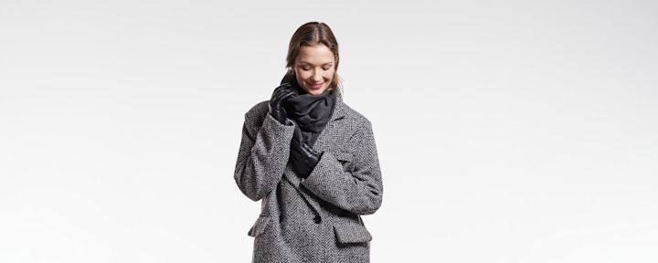 Kvinne smiler og holder skjerf mens hun har på seg grått fiskebensmønstret kåpe og svarte hansker mot hvit bakgrunn.