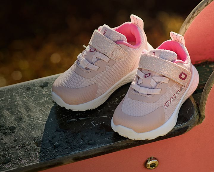 Rosa barnsneakers från Okaki med kardborreband och nätmaterial, placerade på en mörk yta med suddig bakgrund.