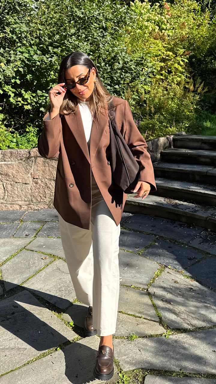 Person i brun kavaj och vita byxor bär bruna lädermokasiner med kraftig sula i en solig trädgårdsmiljö.