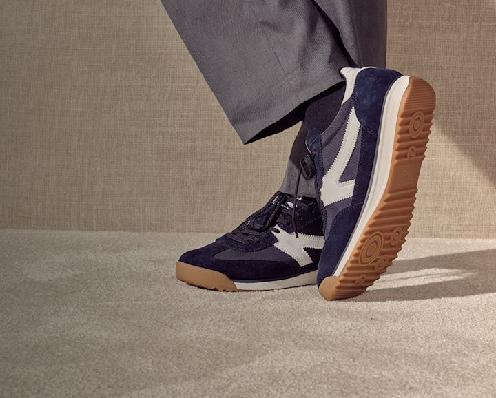 Mørkeblå low profile sneakers med hvite detaljer og gummibunn, brukt med grå bukser mot en nøytral bakgrunn.