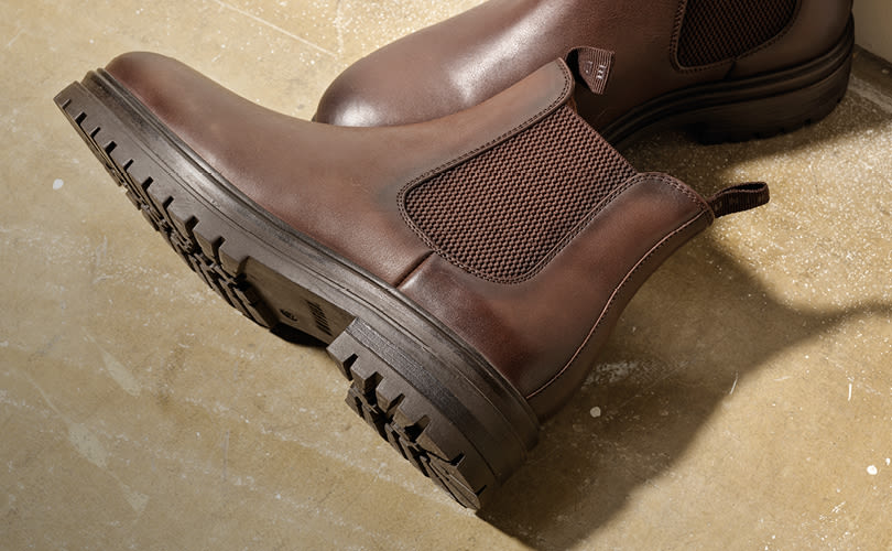 Brune chelsea boots med elastiske sidepaneler og kraftig såle, plassert på et beige gulv.