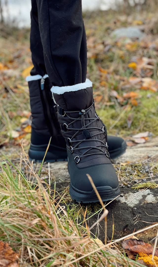 Sorte vinterstøvler med hvit pelskant og snøring på en person som står på en stein i et høstlandskap med gress og løv.