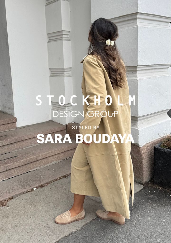 Kvinna med långt, vågigt hår dekorerat med vita blommor går uppför en trappa utanför en byggnad iklädd en beige lång kappa och skor.