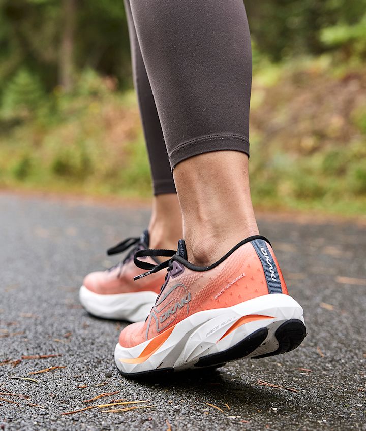 Person i svarta löpartights bär korallrosa Okaki Pace Run med vit sula på en våt asfaltväg i skogsmiljö.