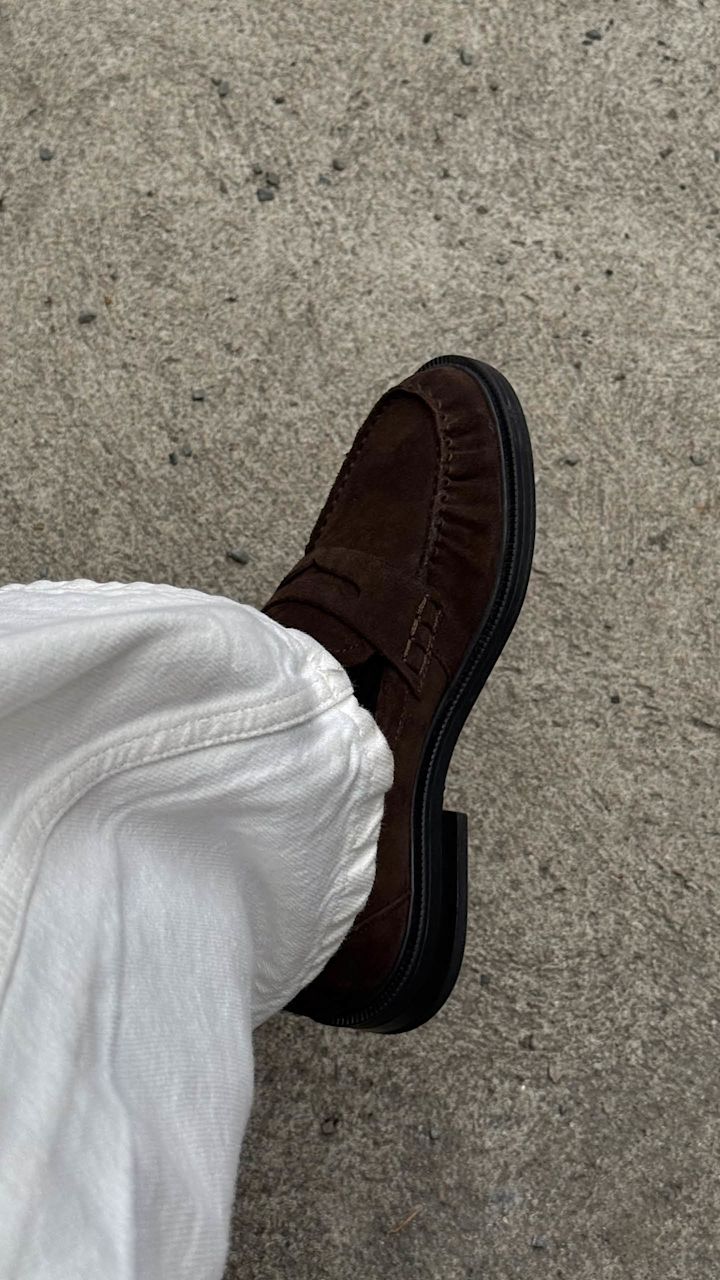Mörkbrun mockasin-loafer med fransar och svart sula, buren med vita jeans på grå betongunderlag.
