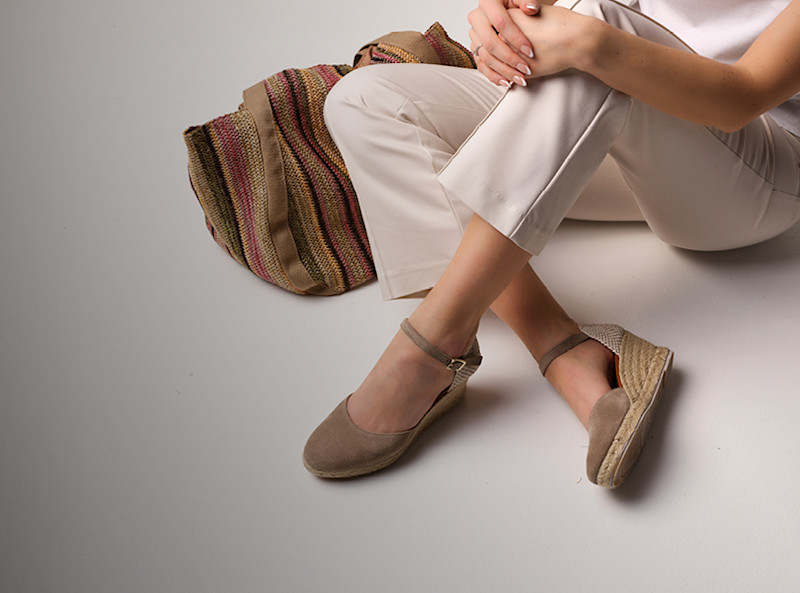 Kvinna i vita byxor med beige mocka espadriller med kilklack och ankelrem, samt en färgglad stickad väska.