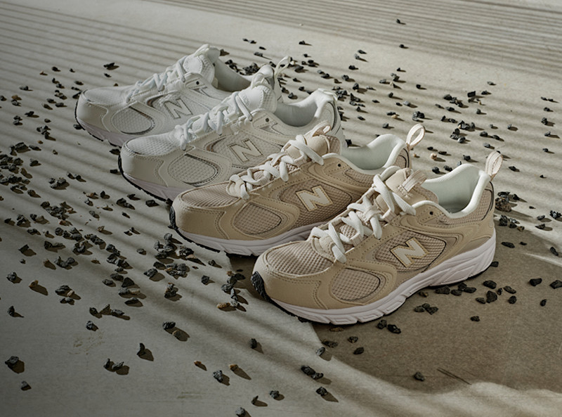 Två par New Balance löparskor i beige och vita nyanser uppställda på en sandyta med små mörka stenar utspridda runtomkring.