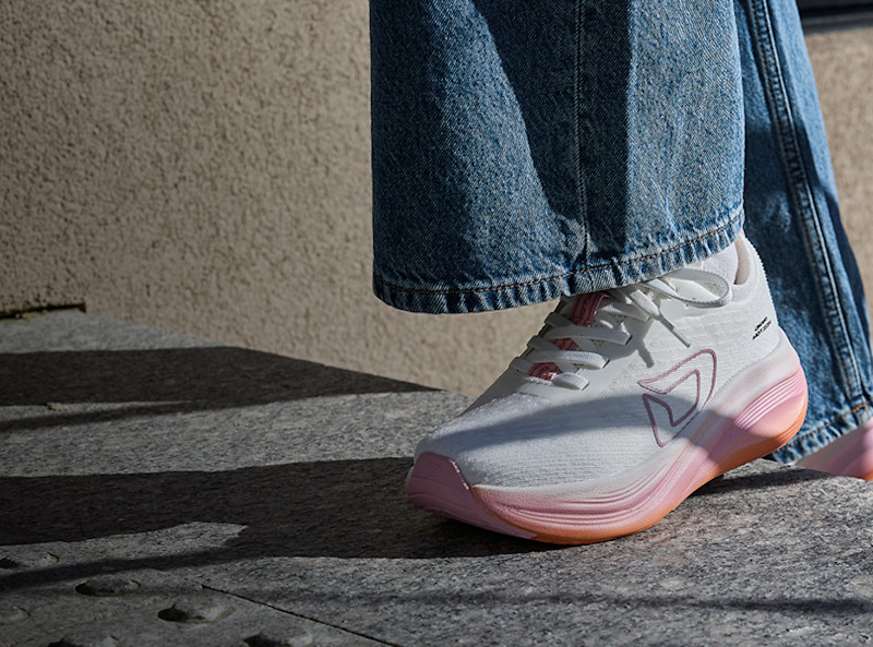 Hvite sneakers fra Okaki med rosa såle og detaljer, brukt med vide jeansbukser, står på en grå steinoverflate.