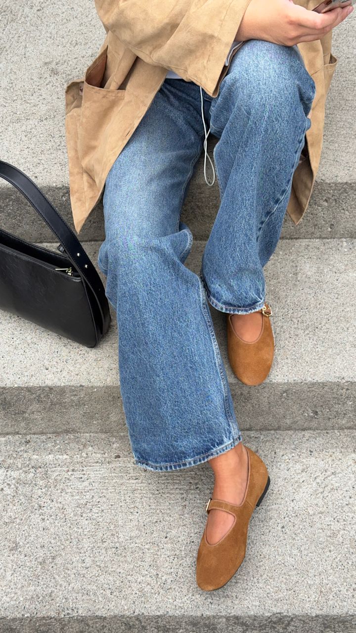 Lisa Alexandra i beige frakk og jeans sitter på trapp med brune semsket ballerinasko og svart veske ved siden av.