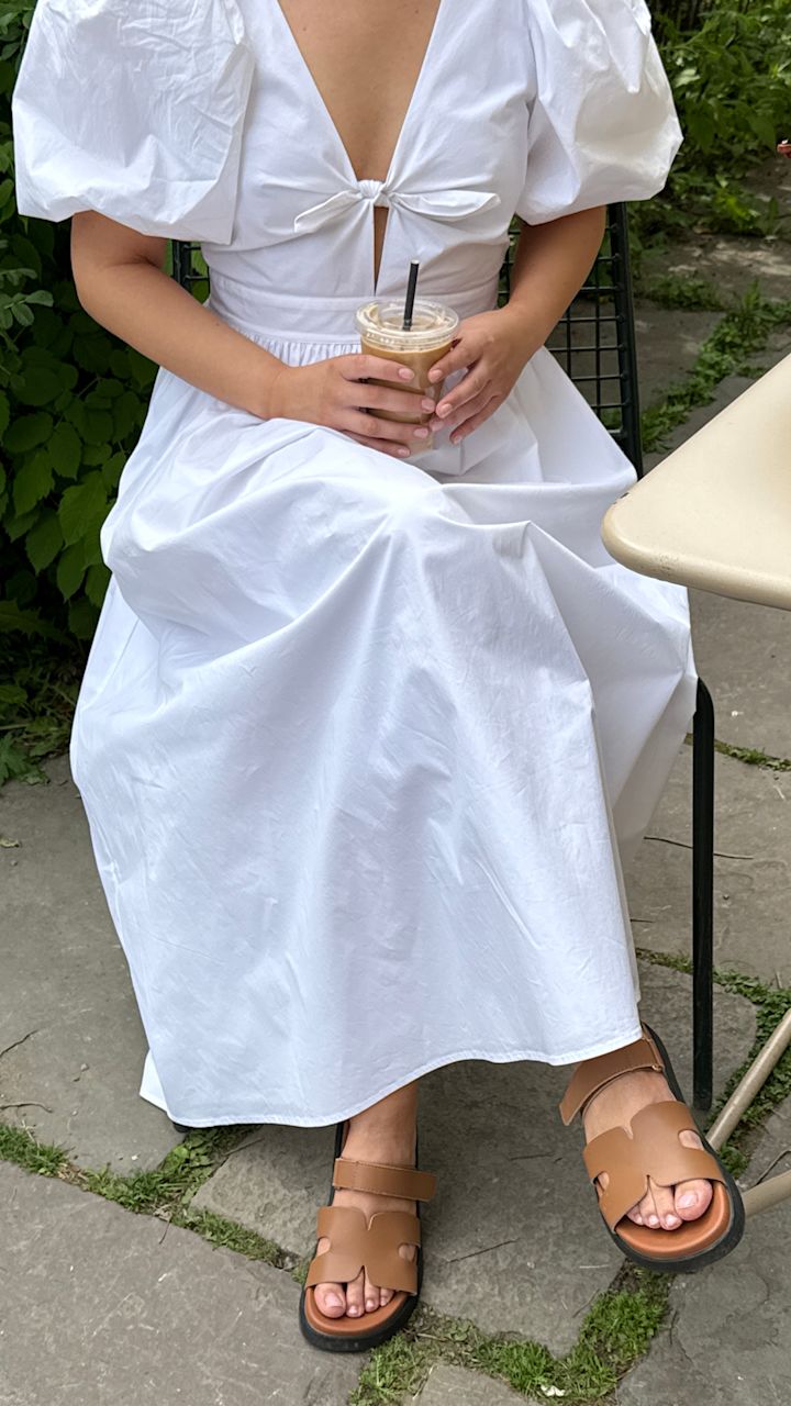 Person sitter utomhus på stol med vit klänning med puffärmar och håller en plastmugg med iskaffe, iklädd bruna sandaler på stenlagd mark.