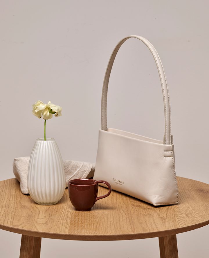 En beige läderväska på ett träbord, bredvid en vit vas med blomma och en brun keramikmugg.