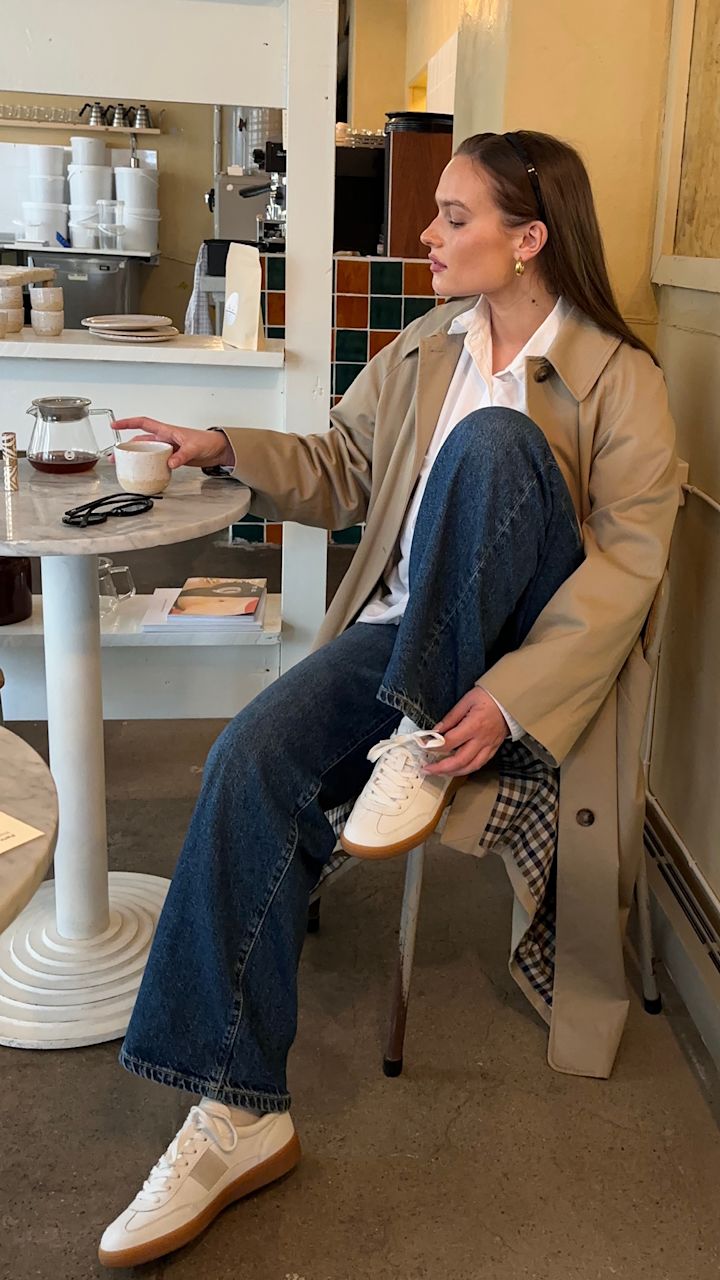 Kvinne i beige trenchcoat og blå jeans sitter på kafé, holder en kopp kaffe med den ene hånden og hviler den andre på benet med hvite sneakers.