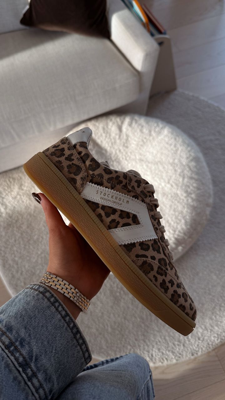 En hand håller en leopardmönstrad sko med vit detalj och texten "STOCKHOLM DESIGN GROUP" i ett ljust rum med vit soffa och rund vit puff i bakgrunden.
