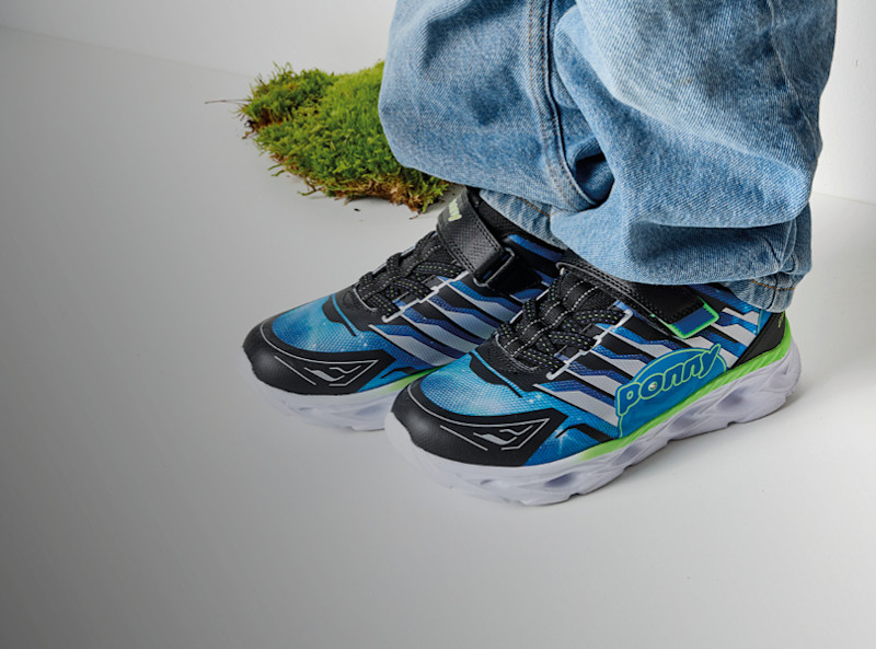 Ponny blinkskor i blå och grön sportig design med kardborreband, burna med jeans på en gräsmatta mot grå bakgrund.