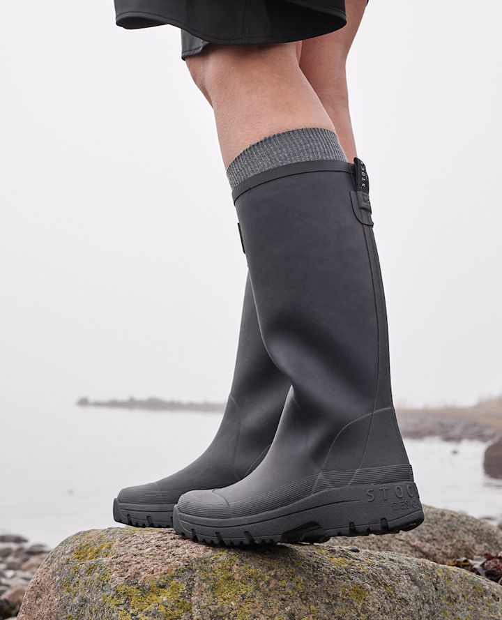 Person står på en stein ved vannet iført høye, mørke gummistøvler og grå ullsokker i et tåkete landskap.