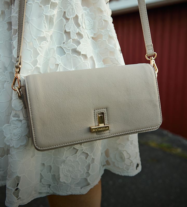 Kvinne i hvit kjole står barbeint på klipper ved sjøen og holder en beige tote bag med svarte håndtak.