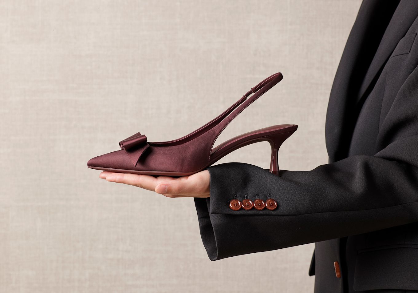 Vinröd slingback-sko med rosett och låg klack hålls i handen av person i svart kostym mot beige bakgrund.