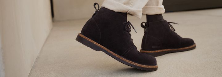 Svarte semsket ankelstøvletter med snøring, korkdetaljer langs sålen og hvite bukseben over mørke sokker.