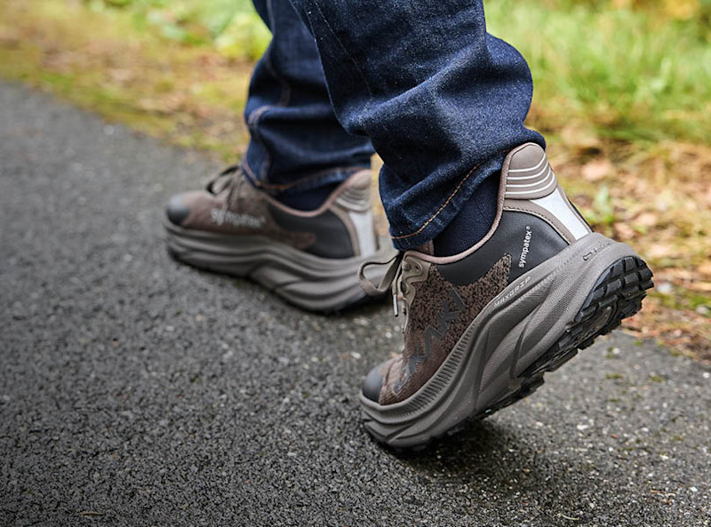Person i blå jeans går på asfalt i brune tursko fra Okaki med grå såle og Sympatex-membran.