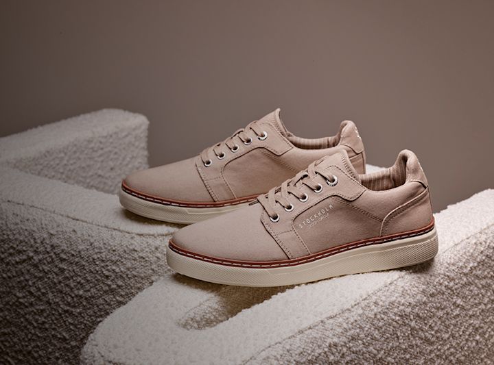 Beige lavskaftede sneakers med hvit såle og brune detaljer, plassert på en lys teksturert overflate.