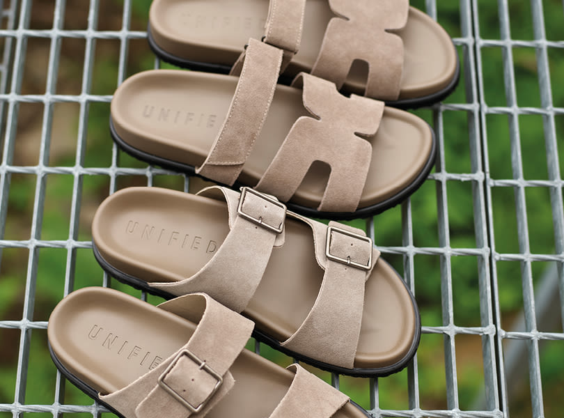 To par beige UNIFIED sandaler i semsket skinn, med ulike remdesign. Plassert på et metallgitter med grønn bakgrunn.