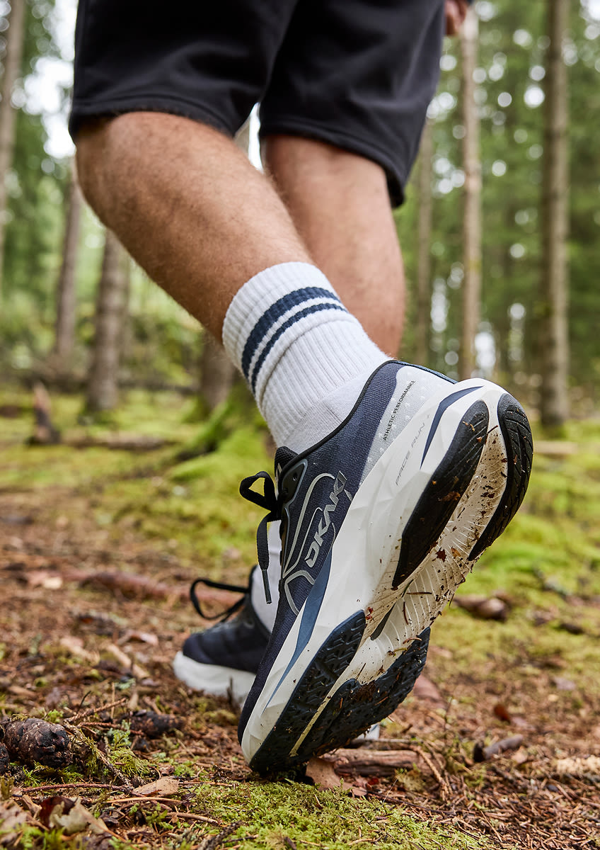 Person i svarta shorts och vita strumpor med blå ränder bär mörkblå och vita  Okaki Pace Run löparskor på en skogsstig.