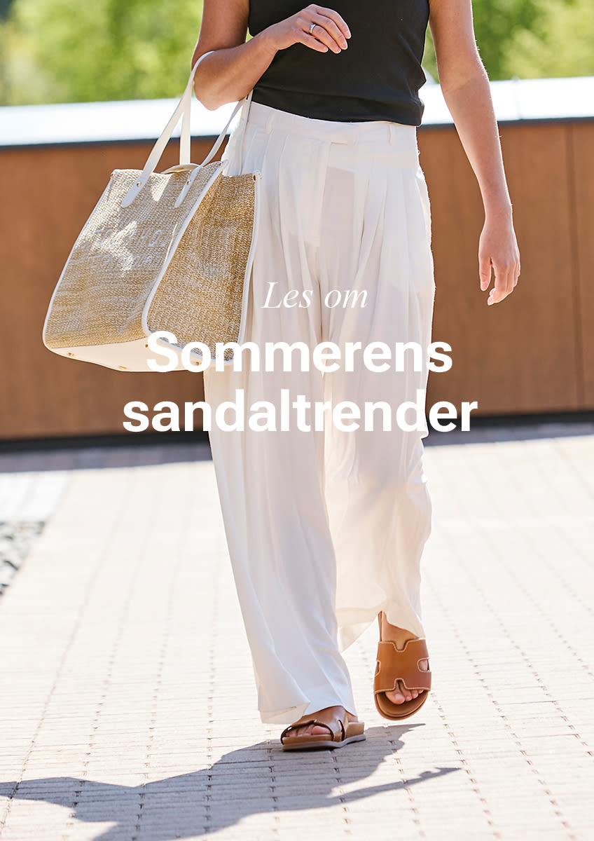 Person i svart topp og hvite bukser bærer stråveske og går med brune, elegante slip-on sandaler i sommerlig stil.