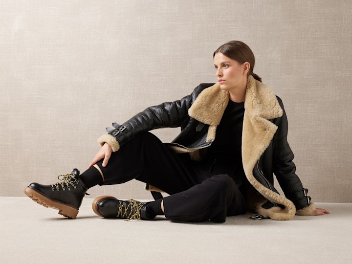 Person i svart läderjacka med beige pälskrage bär svarta vandringskängor med gula skosnören och brun sula mot beige bakgrund.